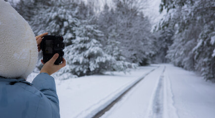 Woman taking a photo of a winter forest. Photographer taking photos of a snowy road winding through a quiet forest surrounded by trees covered in a white blanket, creating a serene winter landscape.
