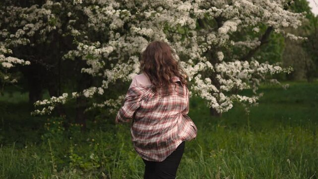 Caucasian woman running on grass towards blossoming tree. Back tracking shot of happy unrecognizable person with flowing hair in spring garden with neutral light.