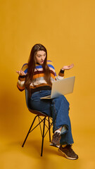 Fototapeta premium Young woman, with confused expression, sitting with laptop, raising hands in frustration, wearing striped sweater, against mustard color studio background. Concept of technology, online shopping.