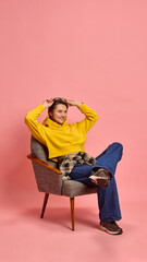 Confident young woman, leaning back in chair, adjusting sunglasses with happy expression against pink studio background. Concept of relaxation, emotions, modern youth, self-assurance.