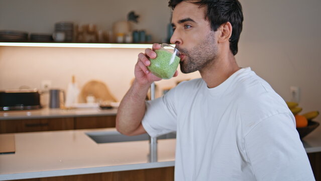 Man drinking green smoothie from glass in modern kitchen closeup. Smiling guy