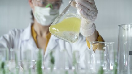 Woman scientist wearing white protective gloves is shaking up a yellow chemical solution inside an...
