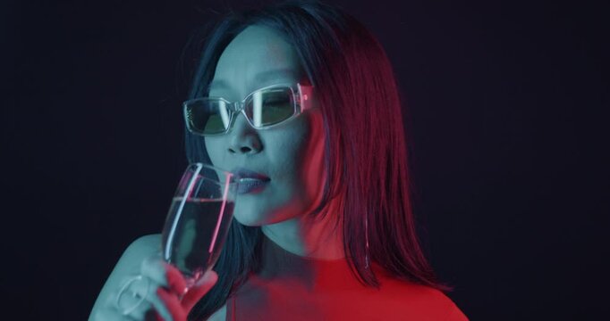 Cheerful Asian lady dancing and drinking champagne in neon light on dark background. Youth culture and celebration concept.