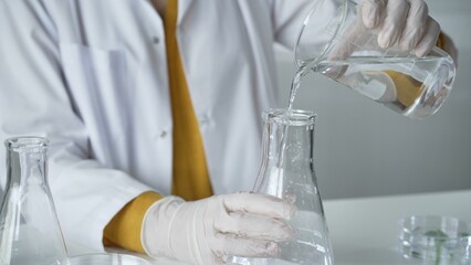 Unknown scientist with a lab coat and white protective gloves is carefully pouring a liquid from a...