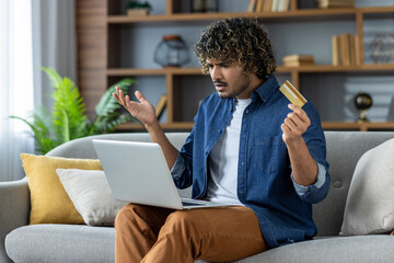 Disappointed deceived man sitting at home on sofa in living room holding bank credit card. Account...