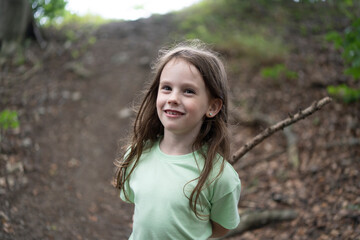 Little cute girl in a t-shirt with a hiking stick on a path in the middle of a summer forest. Concept of children's active travel in nature