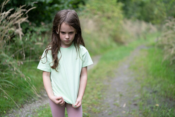Little sad girl in a t-shirt on a forest road in summer. Concept of a lonely child and sadness