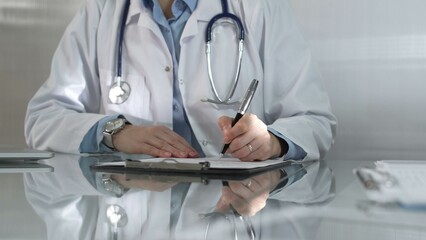 Woman doctor, wearing a blue blouse under a white medical coat, is using a clipboard and taking notes on glass desk in a medical office, close-up. Medicine