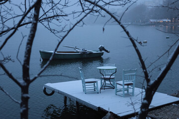 Fototapeta premium snow-covered dock with chairs and a table, overlooking a calm lake, single boat floats nearby
