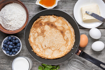 Frying pan with delicious crepes and ingredients for cooking on rustic wooden table. Thin pancakes in a pan with flour, eggs, milk, blueberries and butter. Top view. Delicious breakfast