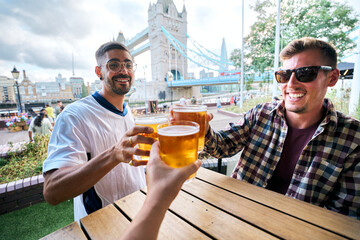 Group of friends enjoying a beer at a pub in london with tower bridge in the background, celebrating and toasting their drinks on a sunny day