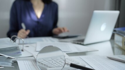 Obraz premium Magnifying glass, white calculator, pen and documents are lying on accountant's desk. Businesswoman wearing blue dress is working with documents on the background. Business and audit concept