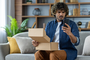 Dissatisfied and upset man sitting at home on the couch holding a phone and a postal parcel box in his hands, mistaken purchase in an online store, dissatisfied buyer client.