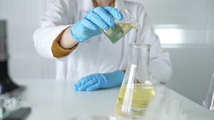 A scientist, wearing a lab coat and blue protective gloves, is pouring a yellow liquid from one beaker to another in laboratory, close up. Medicine and science concepts