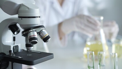 A scientist, wearing a lab coat and pink protective gloves, is pouring a yellow oily liquid from one beaker to another near microscope in laboratory, close up. Medicine and science