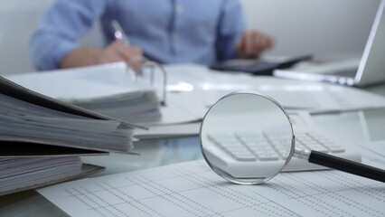 Magnifying glass, calculator, pen and documents are lying on accountant's desk. Businessman wearing blue shirt is working with documents on the background. Business people, audit concept