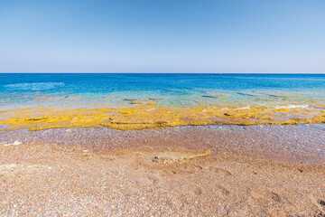 Clear turquoise water of Aegean Sea washing over rocky shore with small pebbles under bright sunlight. Rhodes, Greece