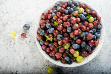 A plate of summer garden berries, gooseberries and blueberries, stands on a gray concrete table.