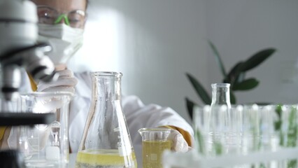 Woman scientist wearing a lab coat, white gloves, and a mask is pouring a yellow oily liquid from one beaker to another in a laboratory setting, close up. Science and medicine
