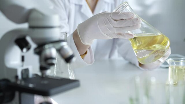 Unrecognizable scientist researcher, wearing white protective gloves and lab coat, is holding a beaker with yellow oil liquid while sitting at the table in laboratory, close up. Medicine and science