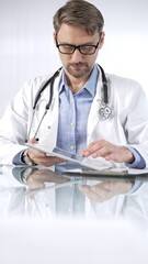 Doctor man in blue shirt under a white coat, glasses and stethoscope is using digital tablet in modern healthcare setting, interacting with patient data, vertical portrait view. Medicine and science
