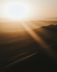 Obraz premium Golden sunlight streams across rolling sand dunes, silhouetting distant mountains in a hazy, serene desert landscape.