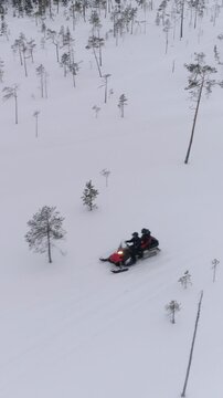 AERIAL CLOSE UP: People driving snowmobile on beautiful white snowy mountain slope exploring Lapland, Finland. Tourists on snowmobile adventure joyride. Travelers snowmobiling through pine tree forest