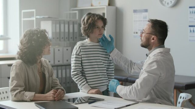 Doctor in white coat and disposable gloves examining throat and lymph nodes of little boy, then talking to his mother sitting beside during medical check-up in pediatric clinic