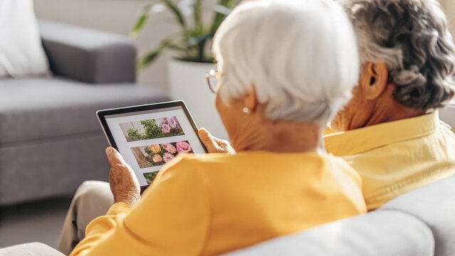 An elderly couple seated together engrossed in a virtual workshop on gardening for health benefits with a tablet positioned between them showing colorful images of plants and