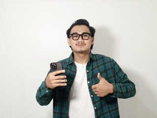Smiling satisfied young Asian man wearing a plaid checkered shirt holding a mobile phone and showing thumbs up sign, recommends apps isolated on white background
