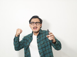 Excited young Asian man wearing a plaid checkered shirt holding a mobile phone and doing a winner gesture isolated on white background