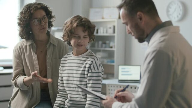 Woman and her little son sitting together on examination table in modern medical office, consulting about treatment with pediatrician