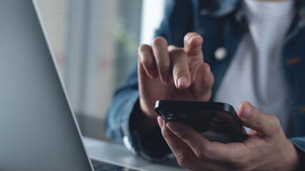 Closeup, business woman sitting at table using mobile smrt phone during working on laptop computer at home office. Young asian woman freelancer  surfing the internet, online working from home