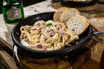 Delicious creamy pasta with ham and grated cheese, served in a rustic black plate with slices of bread. Cozy dining ambiance with wooden table setting