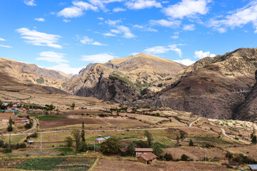 A scenic view of a rural Andean valley featuring traditional houses, farmland, and winding roads, surrounded by majestic mountains under a blue sky