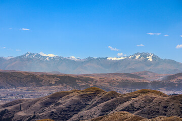 Scenic view of the Andean mountains in Perú, featuring dry grasslands in the foreground, rugged peaks in the midground, and snow-capped mountains under a bright blue sky
