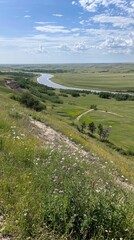 Fototapeta premium Scenic view of a winding river surrounded by lush green fields and wildflowers under a clear sky
