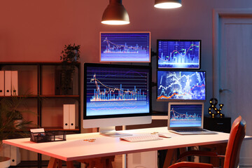 Laptop and computer monitor with stock data on trader's desk in dark office at night