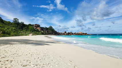 A pristine tropical beach with white sand, turquoise ocean waves, granite rock formations, and lush green vegetation. Seychelles, La Digue.