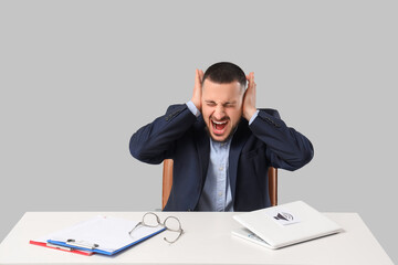 Young businessman suffering from loud noise at table on grey background