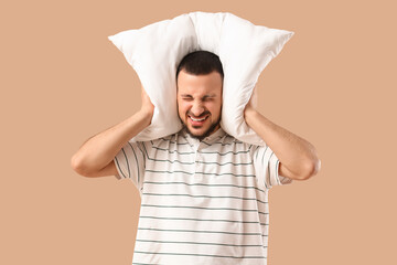 Young man with pillow suffering from loud noise on beige background
