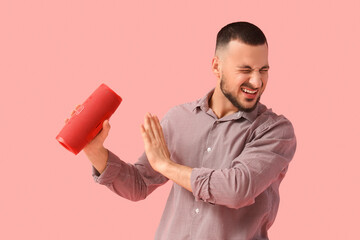 Young man with portable speaker suffering from loud noise on pink background