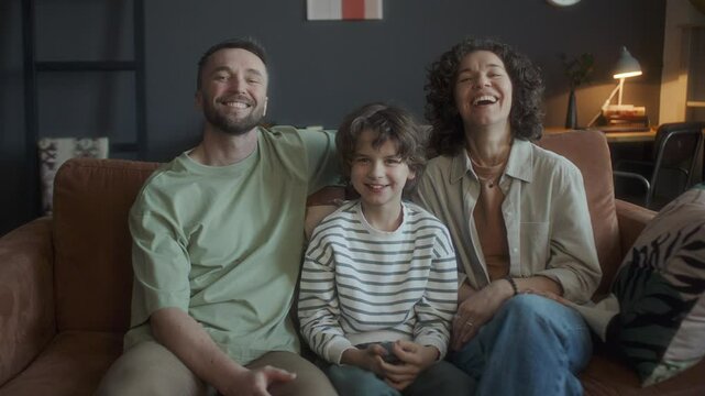 Cheerful parents and their little son sitting together on sofa in cozy living room, watching hilarious TV show, looking into camera and happily laughing in the evening at home