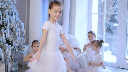 Group of young girls are dressed in white and are dancing in front of a Christmas tree. Scene is festive and joyful, as the girls are dressed in traditional ballet attire