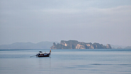 Obraz premium A tranquil boat glides through the calm waters near Koh Hong Island Thailand, with distant limestone formations framing the horizon. The soft light casts an enchanting atmosphere during sunset.