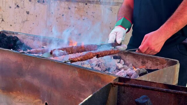 Chef flipping kofta kebab skewers on a charcoal grill in a food booth in the street during daytime