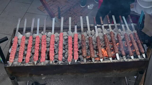 Chef flipping kofta kebab skewers on a charcoal grill in a street food booth during daytime