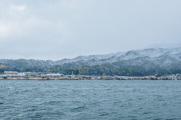 Naklejka premium The winter coastline of Kyoto, Japan, features seaside houses built along the shore and snow-capped mountains in the background. The gloomy weather enhances the desolate atmosphere of winter.