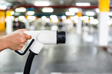 Close-up of a hand holding an electric vehicle charging plug in a parking garage. Symbolizing clean energy, EV infrastructure, sustainability, green technology, and the future of transportation.