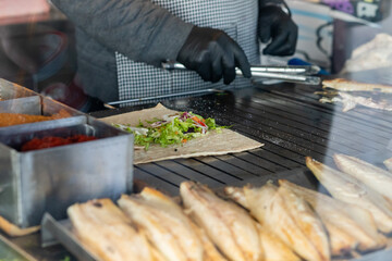 A vendor assembling a wrap with fresh vegetables beside grilled fish on a hot grill, showcasing vibrant street food preparation.
